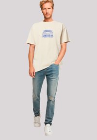Beige Baumwoll-T-Shirt mit blauer Grafik eines Retro-Autos, kombiniert mit hellblauen Jeans aus Denim mit distressed Details und weißen Sneakers.