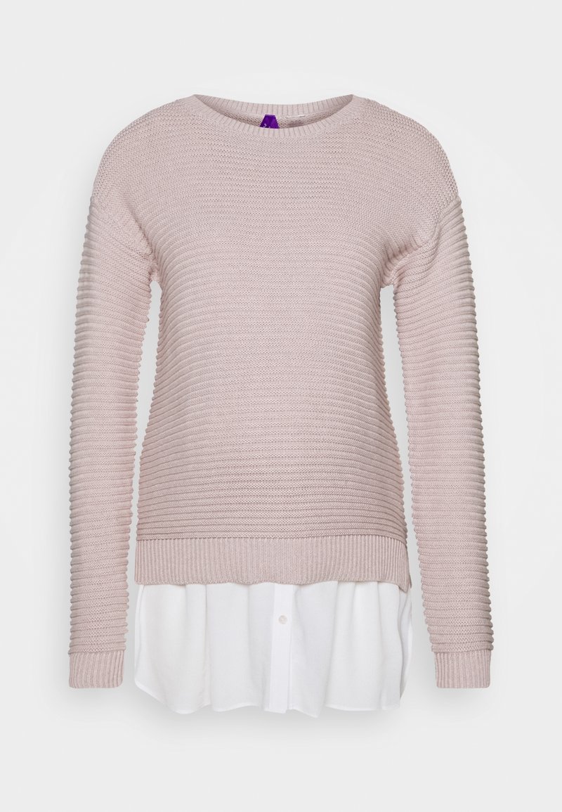 Pull en maille côtelée rose clair superposé sur une chemise blanche boutonnée avec un col arrondi et des manches longues.