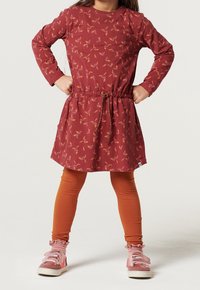 Enfant debout, mains sur les hanches, portant une robe fleurie rouille, des leggings orange brûlé, des chaussettes roses à volants et des baskets roses.