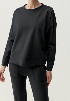 Person iført en ensfarvet sort langærmet sweatshirt og sorte bukser med synlige sømdetaljer mod en neutral baggrund.