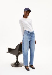 Un haut blanc léger à manches longues, un jean bleu taille haute avec un ourlet effiloché et des ballerines noires, complété par une casquette navy. Une chaise noire en arrière-plan.