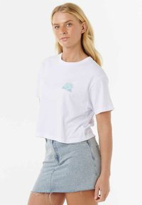 Weißes, kurzgeschnittenes T-Shirt aus Baumwolle mit einem kleinen türkisfarbenen Grafikdruck mit dem Text "Rip Curl" auf der linken Brustseite. Kombiniert mit einem Jeansrock.