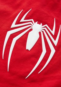 Rødt stof med et stort hvidt spider-emblem med stiliserede ben og krop. Designet har en glat, hævet tekstur mod materialet.
