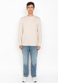 Camicia in cotone beige a maniche lunghe con colletto rotondo, abbinata a jeans in denim azzurro chiaro e sneakers grigie, in piedi davanti a uno sfondo bianco.