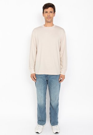 Beige langærmet bomulds skjorte med rund hals, parret med lyseblå denimjeans og grå sneakers, stående mod en hvid baggrund.