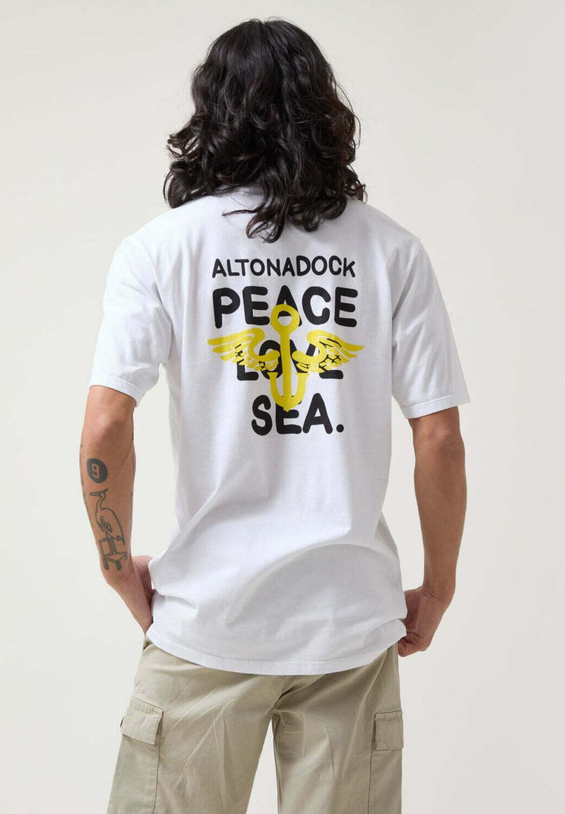 Camiseta de algodón blanca con un gráfico en negro y amarillo en la parte posterior que presenta el texto "ALTONADOCK PAZ AMOR MAR" y un diseño de ancla con alas.
