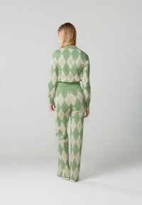 Hátát fordító szőke hajú nő zöld és bézs színű, argyle mintás pulóverben és hozzá illő nadrágban, egyszerű háttér előtt.