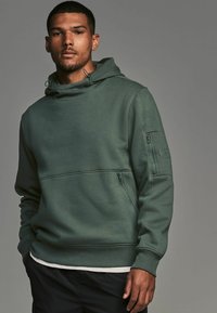 Sudadera con capucha de tejido verde con un bolsillo de cremallera en la manga izquierda, puños acanalados y un acabado suave y texturizado.