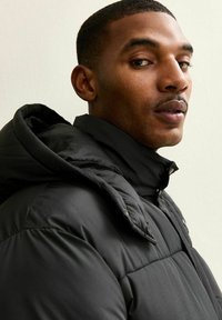 Chaqueta acolchada negra con cuello alto y costuras visibles. Textura suave y acabado blando, con cierre de cremallera y capucha.