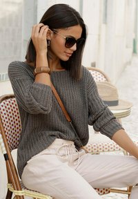 Grauer Strickpullover mit aufgerollten Ärmeln, kombiniert mit hellbeigen, schmalen Hosen. Eine braune Umhängetasche und schwarze Sonnenbrillen sorgen für Kontrast.