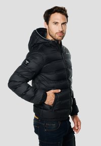 Schwarze Steppjacke mit Kapuze, ausgestattet mit einem Reißverschluss, horizontalen Steppnähten und elastischen Bündchen. Getragen zu blauen Jeans.