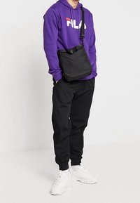 Persona con un hoodie viola FILA, pantaloni jogger neri, sneakers bianche e una borsa a tracolla nera, in piedi davanti a uno sfondo bianco semplice.