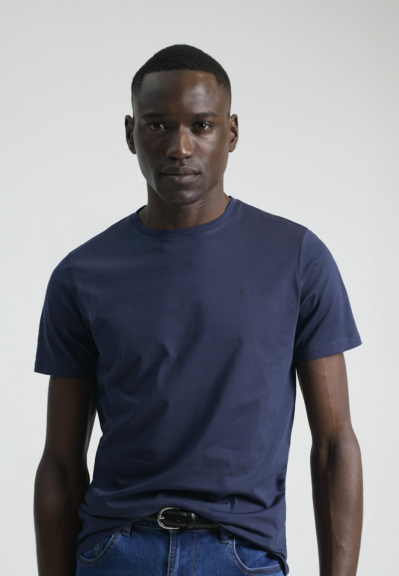 Camiseta de algodón azul marino con cuello redondo y mangas cortas, con un pequeño logo en el pecho. La lleva puesta con unos jeans de mezclilla y un cinturón negro.