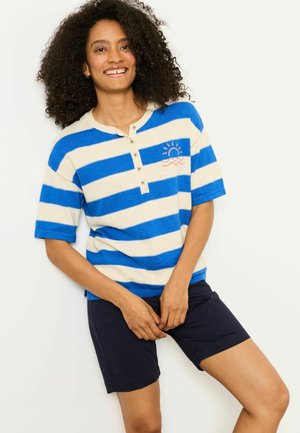 Frau mit lockigem Haar, die lächelt, trägt ein blau-weiß gestreiftes Kurzarmoberteil mit Sonnenstickerei und marineblaue Shorts, steht vor einer weißen Wand.