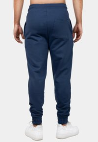 Navy blauwe trainingsbroek met een aansluitend ontwerp, ribgebreide manchetten en een enkele achterzak. Gemaakt van zacht, gemeleerd stof. Witte sneakers zichtbaar.