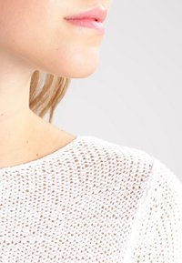 Detailní záběr dolní části obličeje, krku a ramene osoby oblečené v bílém texturovaném pleteném topu na pozadí jednobarevné světlé barvy.