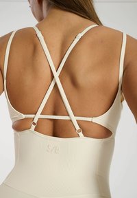 Ribbede beige body med justerbare krydsbare stropper. Har logodetaljer og en glat tekstur. Minimalistisk design.