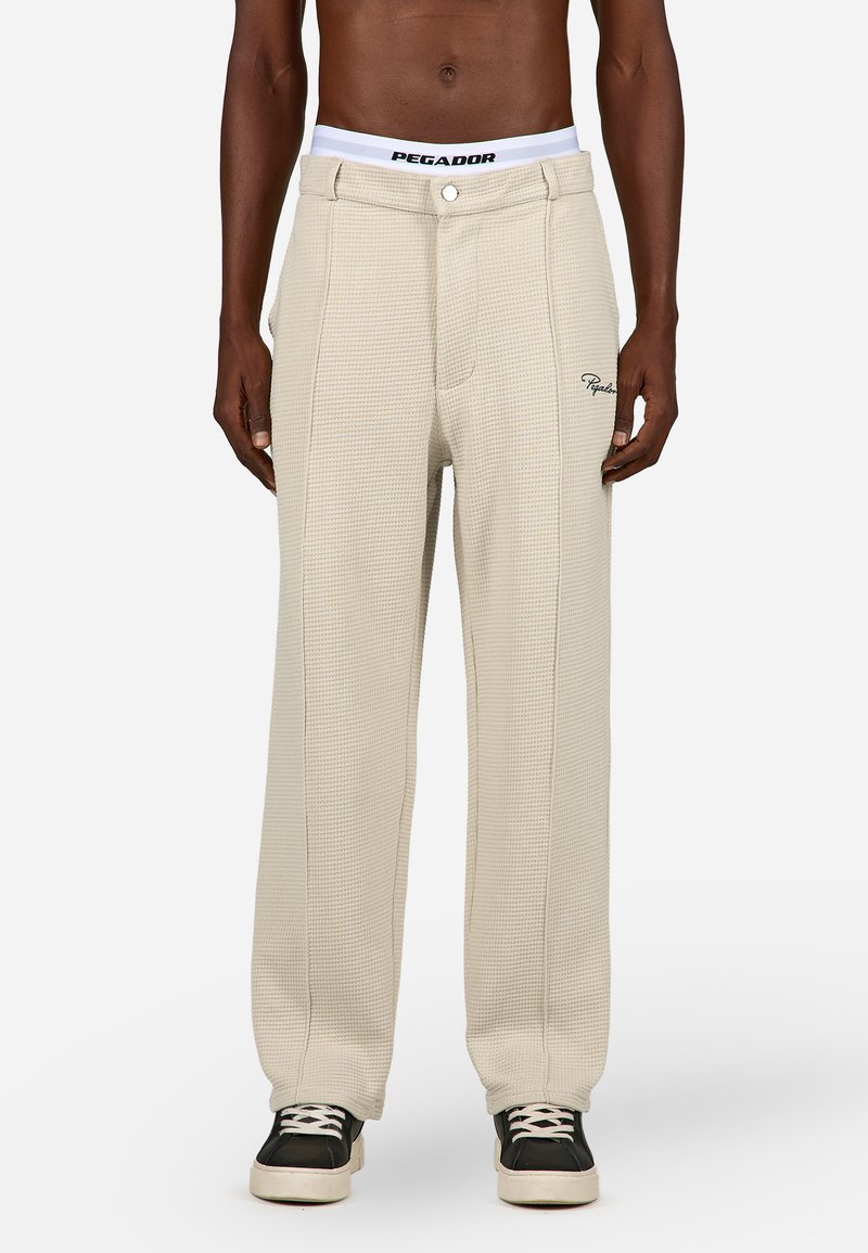 Bež hlače z žametno teksturo in sproščenim krojem, z belim pasom, na katerem je logo "PEGADOR" ter vezenina na strani.