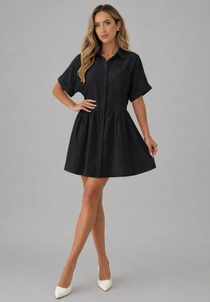Femme aux longs cheveux blonds portant une robe courte noire boutonnée et des talons blancs, debout avec une main sur la hanche et l'autre tenant la robe.