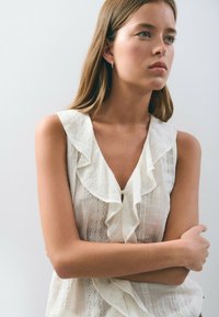 Blusa blanca sin mangas con un escote con volantes, que presenta detalles de encaje en la parte delantera. Tejido ligero y texturizado con un corte holgado.