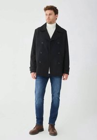 Mann in schwarzem, doppelt geknöpftem Mantel, weißem Rollkragenpullover, blauen Jeans und braunen Stiefeln, der vor einem einfarbigen Hintergrund steht.