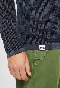 Navy gestrickter Pullover mit strukturiertem Muster, ausgestattet mit einem gerippten Bündchen und Saum. Dazu grüne Hosen mit seitlichen Taschen.