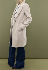 Beige wool-blend coat with a single button, notch lapels, and side pockets, worn over a white and black striped top and flared jeans.