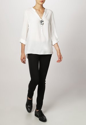 Kobieta ubrana w białą bluzkę z długim rękawem, czarne spodnie i czarne oksfordy z ćwiekami, idąca na jednolitym jasnym tle.