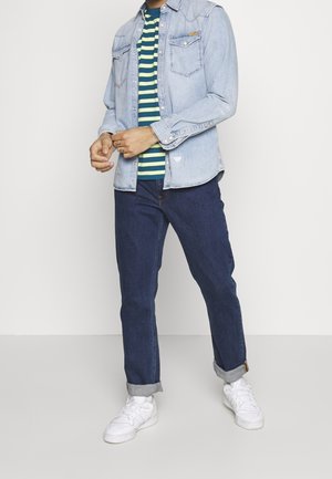 Lichtblauwe denimoverhemd, gelaagd over een groen en marineblauw gestreept t-shirt. Gecombineerd met donkerblauwe opgerolde jeans en witte sneakers.