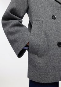 Szary płaszcz z wełny z domieszkami o teksturowanej powierzchni, podwójnie zapinany, z dwoma przednimi kieszeniami i czarnymi guzikami. Rękaw lekko odsłania niebieską koszulę znajdującą się pod spodem.