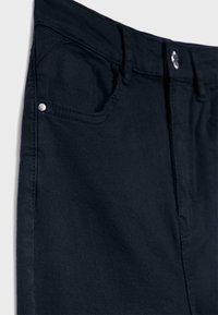 Detailný záber na čierne rifle z denimu zobrazujúci predný vrecko, očko na opasok, stehy a detaily strieborného gombíka.