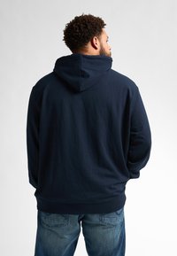 Námořnická modrá mikina s kapucí, vyrobená z měkké látky. Obsahuje žebrované manžety a lem. V kombinaci s světle modrými džínami.