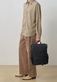 Camicia button-up a maniche lunghe color tan, pantaloni in velluto corduroy beige e scarpe chiare. Zaino nero con doppie spalline e tasche laterali.