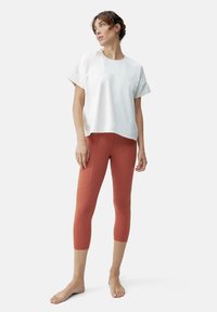 Balta trumpa rankovėmis marškinėliai su laisvu kirpimu ir apvaliomis iškirptomis, derinami su rūdžių spalvos Capri tipo leginsais su šoninėmis siūlėmis ir kišenėmis.