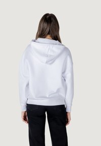 Sudadera blanca con una gran capucha y hombros caídos. Textura suave, puños y dobladillo acanalados. Llevada con pantalones negros.