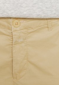 Beige bomullshängselbyxor med knäppning, med en slät yta, bältesöglor och subtila sömnadsdetaljer nära fickan.