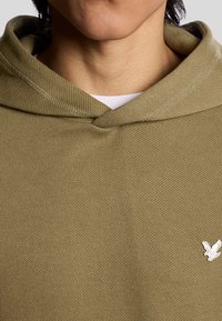 Olijfgroene hoodie met een gestructureerde weving, voorzien van een kangoeroezak, trekkoordcapuchon en subtiele witte logo detail op de borst.
