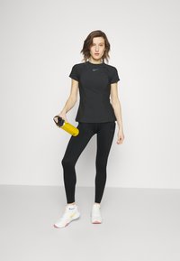 Črna kratka športna majica z sivimi detajli, črne leggings hlače in bele superge z rumenimi in roza detajli; drži rumeno plastenko.