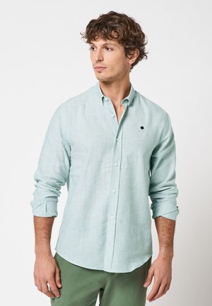 Mann mit lockigem Haar, der ein hellblaues Hemd mit hochgekrempelten Ärmeln und grüne Hose trägt, schaut seitlich vor einem einfarbigen Hintergrund.