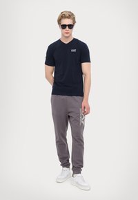 Námornícky bavlnený tričko s výstrihom do V a malým logom, v kombinácii so sivými teplákmi s bielou potlačou a bielymi teniskami.