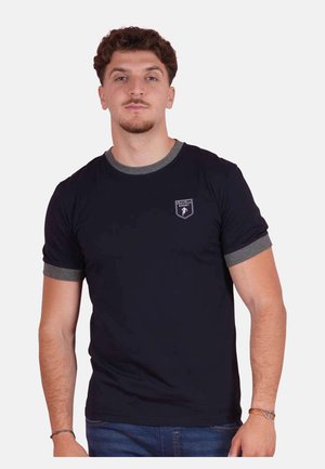 Jeune homme portant un t-shirt noir avec des bordures grises aux manches et au col, debout devant un fond uni clair.