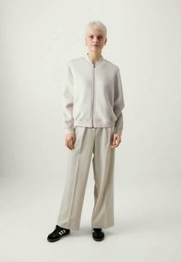 Gai pelēks kapucis ar rāvējslēdzēju, valkāts virs krēmkrāsas platām biksēm. Melni sporta apavi ar baltām svītrām noslēdz kopskatu.