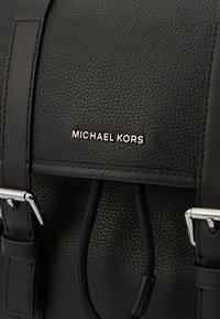 Rucsac din piele neagră cu suprafață texturată, dotat cu accesorii din metal argintiu, bretele ajustabile și un logo proeminent pe față.