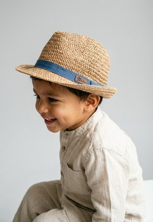 Smiling young child wearing a beige buttoned outfit and a woven straw hat with a blue band, facing left against a plain background.