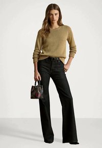 Eine Frau steht in einem beigen Pullover und schwarzen weit geschnittenen Jeans und hält eine kleine schwarze Handtasche mit bunten Designs.