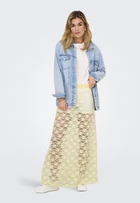 Jeans jakna čez belo majico, v kombinaciji s prosojno rumeno čipkastimi krili z cvetličnim vzorcem; model nosi bele superge.
