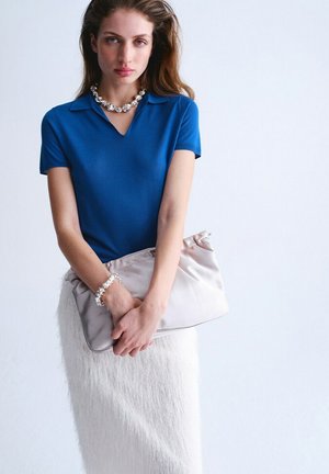 Mujer con blusa azul de manga corta y falda blanca con textura que sostiene un bolso clutch grande de color gris claro, lleva un collar grueso de plata y una pulsera, sobre un fondo liso.
