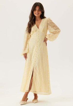 Kvinna som ler, klädd i en lång, ljusgul spetsklänning med ballongärmar, V-ringning, knappar fram och en slits framtill, stående mot en ljus bakgrund.