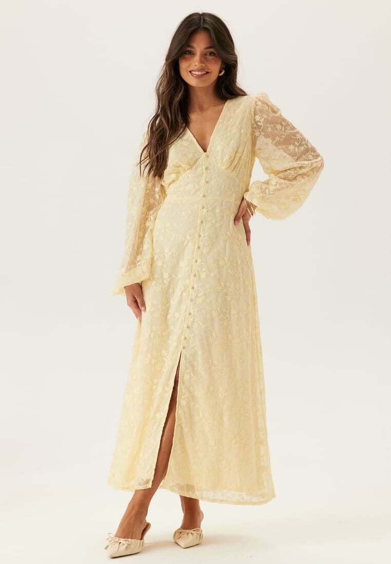 Žena se usmívá, nosí dlouhé, světle žluté krajkové šaty s balónovými rukávy, výstřihem do V, předními knoflíky a předním rozparkem, stojí na světlém pozadí.