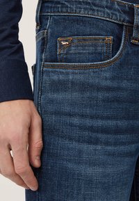 Jeans in denim blu scuro, caratterizzati da un design classico, evidenti cuciture arancioni e un piccolo dettaglio decorativo nella tasca.